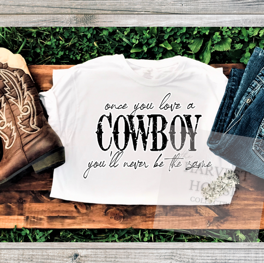 White t-shirt with 'once you love a cowboy you'll never be the same' text, paired with cowboy boots and jeans on a wooden surface.