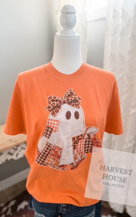 An orange short-sleeve graphic T-shirt displayed on a white mannequin torso, paired with blue jeans. The shirt features a patchwork-style ghost design with leopard-print and plaid patterns.